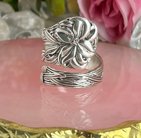 Close-up of an intricately detailed silver floral spoon ring on a pink decorative surface with roses in the background.