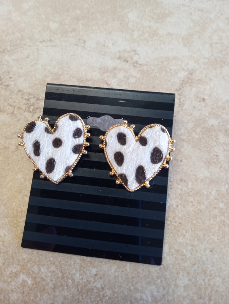 Pair of heart-shaped earrings with white cowhide and black spots, bordered in gold trim on a black striped earring card