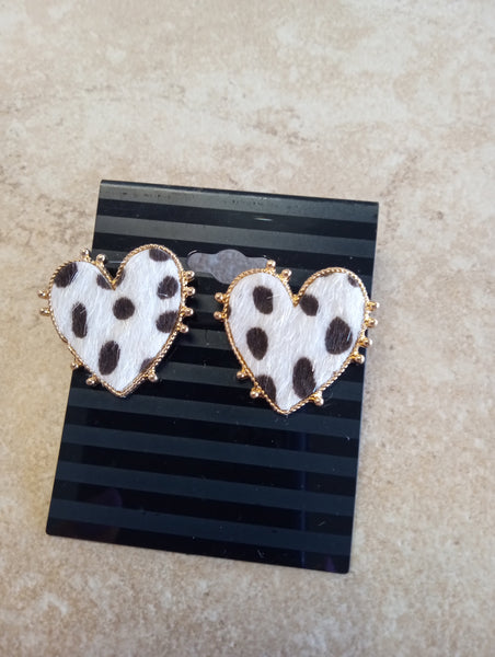 Pair of heart-shaped earrings with white cowhide and black spots, bordered in gold trim on a black striped earring card