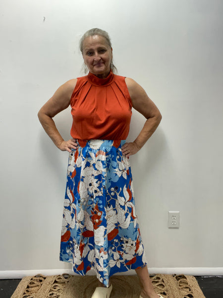 Woman wearing a rust sleeveless top and a bold blue, red, and white floral midi skirt standing against a plain wall.