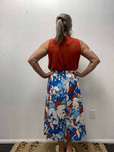 Woman wearing a rust sleeveless top and a bold blue, red, and white floral midi skirt standing against a plain wall.