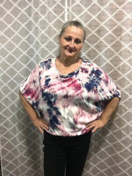 Woman wearing pink, blue and white tie-dye oversized top with ocean wave pattern, hands on hips, smiling, against white background