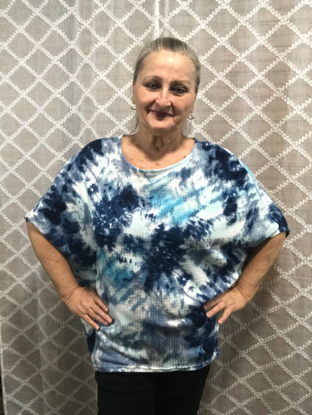 Woman wearing blue and white tie-dye oversized top with ocean wave pattern, hands on hips, smiling, against white background