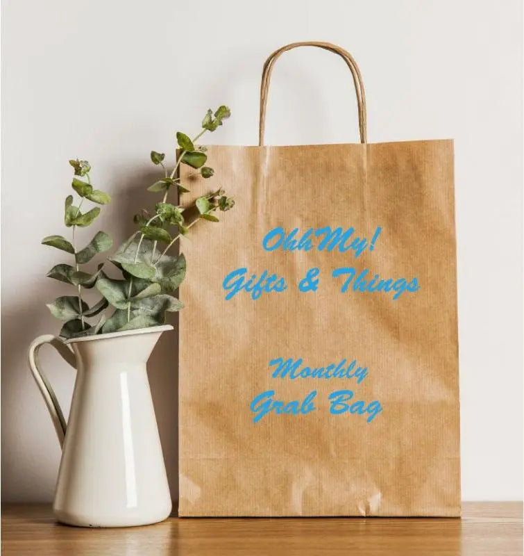 Brown kraft paper gift bag with blue text reading “OhhMy! Gifts & Things Monthly Grab Bag” next to a white pitcher with eucalyptus branches, sitting on a wooden surface.