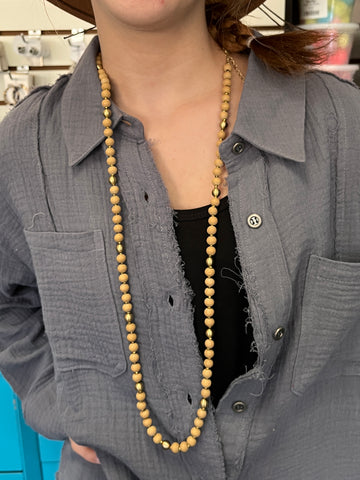 A long beaded necklace featuring natural wood beads accented with gold spacers, styled over a textured gray button-up shirt.