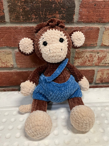Crocheted plush monkey toy wearing blue overalls sitting against a brick wall background