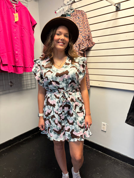 A young woman wearing a pastel camouflage ruffle dress with a belt, paired with white sneakers and a brown wide-brim hat, styled for a casual boutique look.