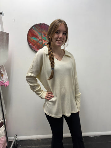 Young woman wearing a cream waffle knit long sleeve pullover top with a soft V-neck, paired with black pants, posing in a boutique setting.