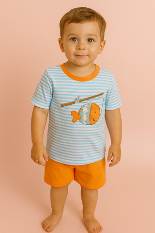 Toddler boy wearing a light blue and white striped t-shirt with a fishing graphic and orange shorts, standing barefoot on a peach background.