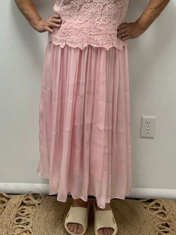 A woman wearing a blush pink pleated maxi skirt with a pink crochet lace top, standing indoors with beige shoes on