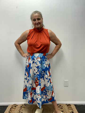 Woman wearing a rust sleeveless top and a bold blue, red, and white floral midi skirt standing against a plain wall.
