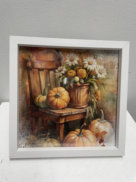 Framed artwork of pumpkins and flowers on a wooden chair against a gray background