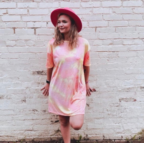 Peach and pink tie dye short sleeve t-shirt dress worn with a wide brim pink hat against a white brick wall