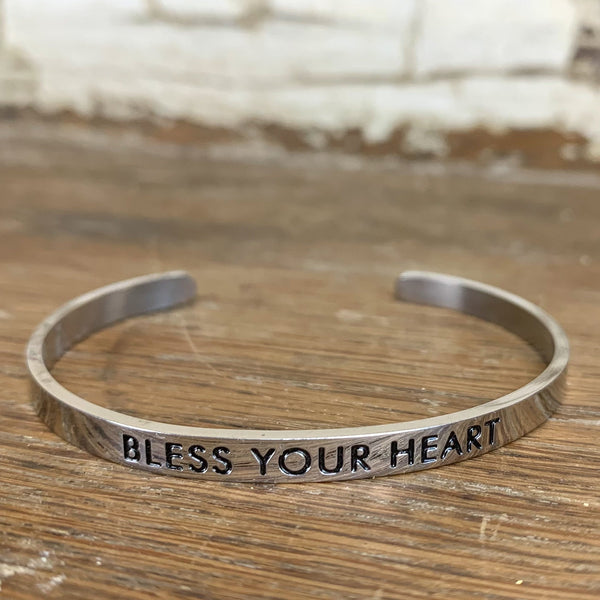 A silver cuff bracelet engraved with the phrase “Bless Your Heart” resting on a rustic wooden surface.