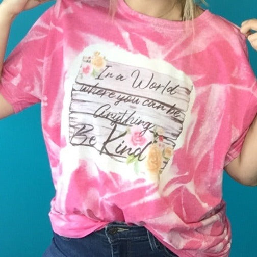 Pink sublimated shirt that says "In a world where you can be anything Be kind" on a wooden background with flowers